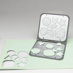 May include: A grey metal die-cutting machine with a variety of shapes and designs. The machine is open and the die-cuts are visible. The die-cuts are in the shape of a flower, arrow, star, button, triangle, and a circle. The die-cuts are made of a light green material.