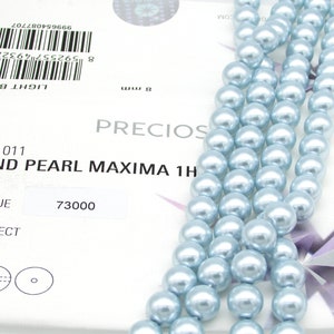 May include: A strand of light blue round pearls, 8mm in size. The pearls are strung on a white thread and are arranged in a straight line. The pearls are smooth and have a high luster. The image also shows a product label with the text "ROUND PEARL MAXIMA 1H" and "LIGHT BLUE".