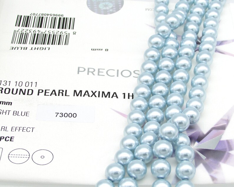 May include: A strand of light blue round pearls, 8mm in size. The pearls are strung on a white thread and are arranged in a straight line. The pearls are smooth and have a high luster. The image also shows a product label with the text "ROUND PEARL MAXIMA 1H" and "LIGHT BLUE".