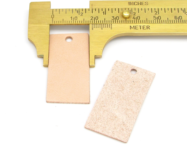 May include: Two rectangular leather tags with a hole punched in the top. The tags are measured with a brass caliper, showing the larger tag is 1.5 inches wide.