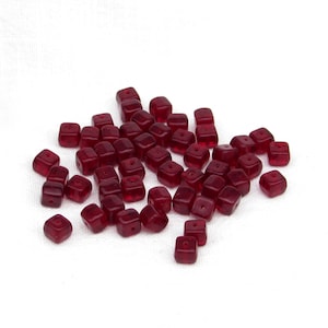 May include: A collection of small, translucent, deep red cube-shaped beads. Each bead features a single, centered hole drilled through the middle. The beads are scattered in a loose pile, displaying a uniform dark crimson color and a smooth, polished finish.