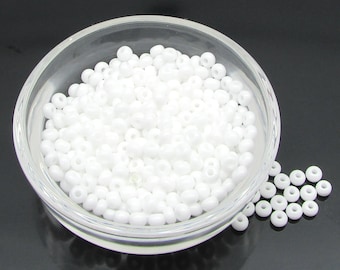 May include: A bowl filled with white seed beads. The beads are small and round, and they are arranged in a random pattern. There are also a few beads scattered on the surface next to the bowl.