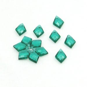 May include: A group of ten emerald green diamond-shaped rhinestones. The rhinestones are scattered on a white surface.