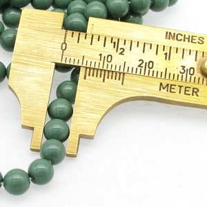 Pu&ograve; includere: Primo piano di una perla verde misurata con un calibro in ottone. Il calibro indica "INCHES" e "METER".