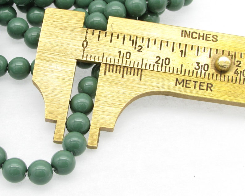 Pu&ograve; includere: Primo piano di una perla verde misurata con un calibro in ottone. Il calibro indica "INCHES" e "METER".