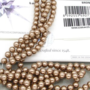 May include: A strand of bronze-colored glass beads. The beads are round and have a smooth, shiny finish. The beads are strung on a clear thread. The beads are approximately 6 mm in diameter. The image also includes a label that reads "BRONZE" and "crafted since 1548."