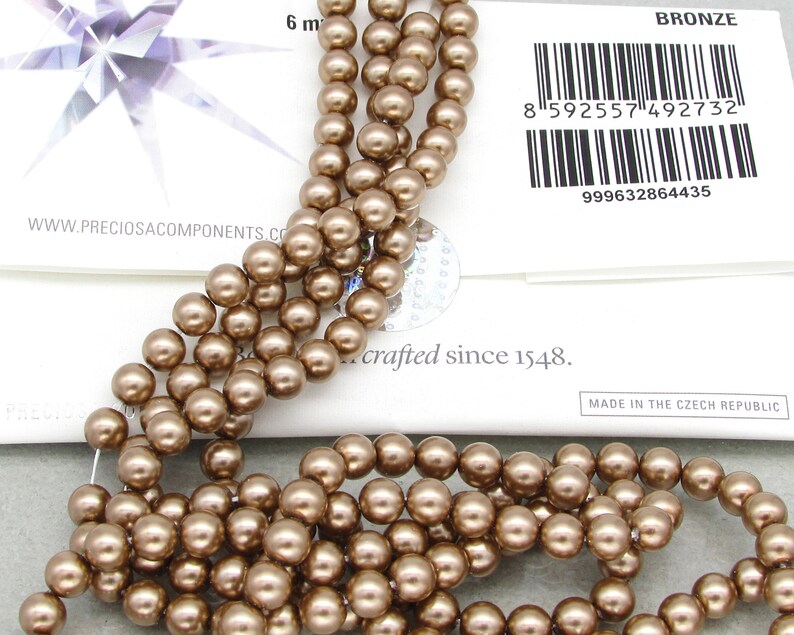 May include: A strand of bronze-colored glass beads. The beads are round and have a smooth, shiny finish. The beads are strung on a clear thread. The beads are approximately 6 mm in diameter. The image also includes a label that reads "BRONZE" and "crafted since 1548."