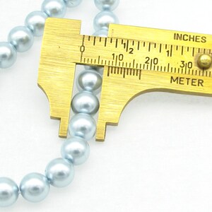 May include: A close-up of a strand of light blue pearl beads being measured with a brass ruler. The ruler shows measurements in inches and centimeters.