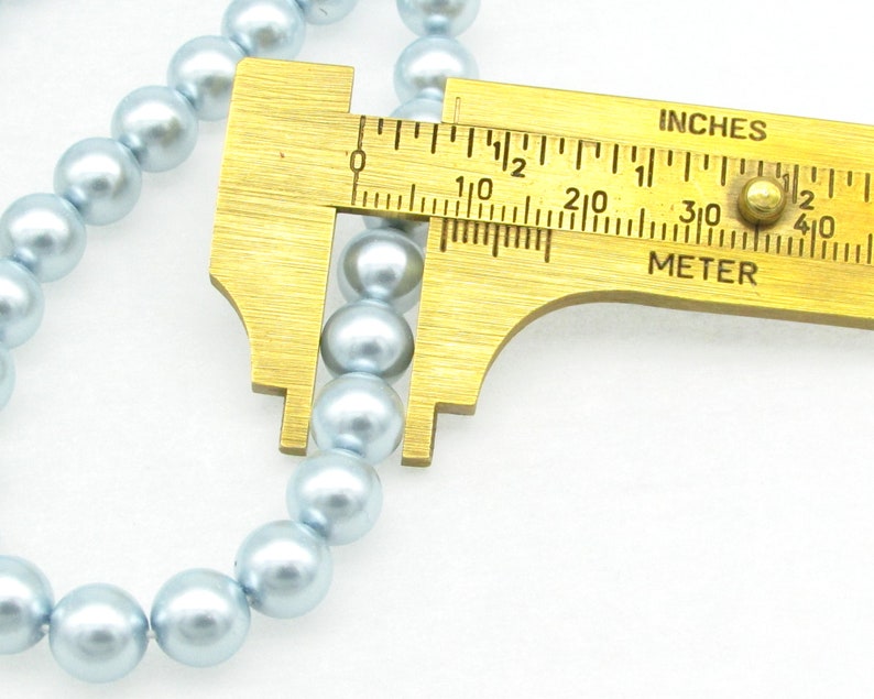 May include: A close-up of a strand of light blue pearl beads being measured with a brass ruler. The ruler shows measurements in inches and centimeters.