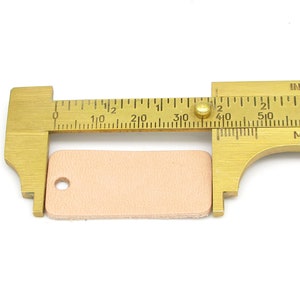 May include: A brass measuring tool with a ruler marked in inches and centimeters. A small, light pink leather rectangle is being measured, measuring approximately 1 1/2 inches in length.