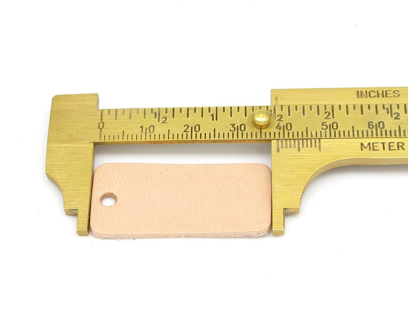 May include: A brass measuring tool with a ruler marked in inches and centimeters. A small, light pink leather rectangle is being measured, measuring approximately 1 1/2 inches in length.