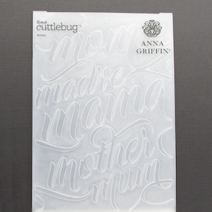 May include: A clear plastic sheet with the words "mom", "madre", "mamma", "mother", and "mum" written in a cursive font. The sheet is for use with a Cricut cutting machine. The text "Cricut cuttlebug" and "ANNA GRIFFIN" is also visible on the sheet.
