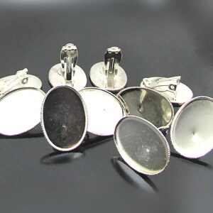 May include: Silver tone earring blanks with a clip-on closure and oval-shaped glass cabochon settings. The settings have a beaded edge.
