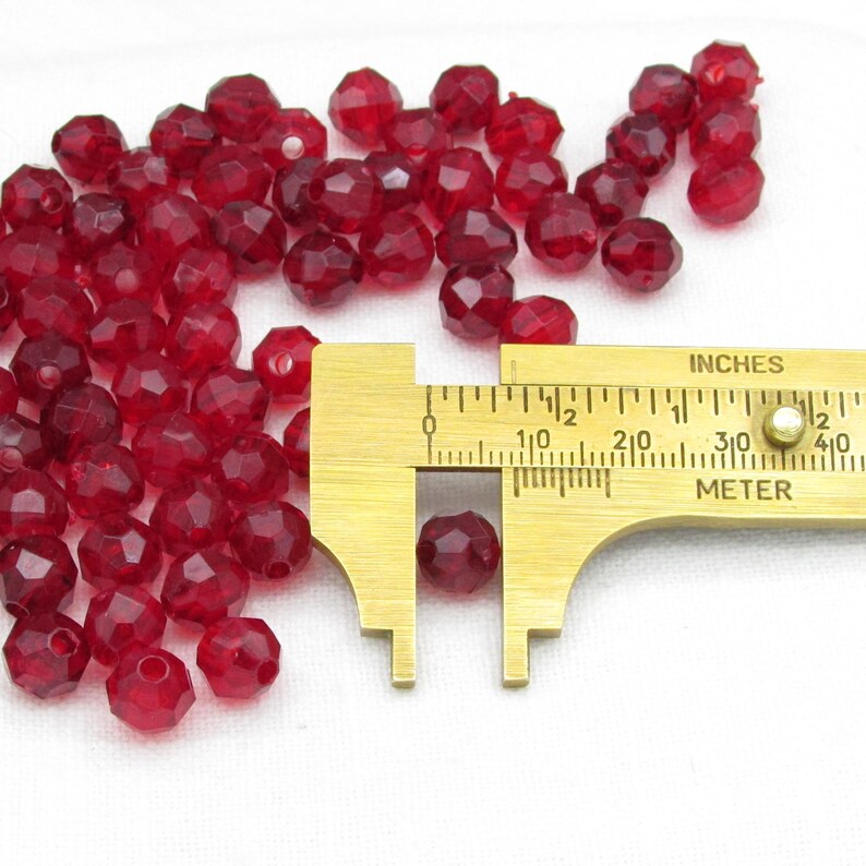May include: A collection of faceted, translucent red beads with a golden measuring caliper. The caliper shows a bead measuring approximately 0.3 inches in diameter. The beads are scattered on a white surface.