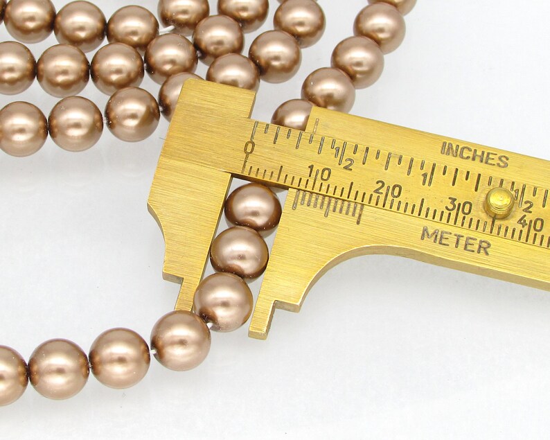 May include: A close-up of a strand of brown pearl beads being measured with a caliper. The caliper reads 1/2 inch or 1.27 cm.