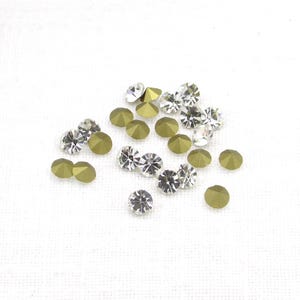 May include: A collection of small, sparkling rhinestones in two colors: clear and olive green. The clear rhinestones have multiple facets, reflecting light. The olive green rhinestones have a smooth, rounded surface. These decorative embellishments are scattered on a white background.