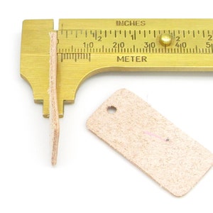 May include: A close-up of a leather tag with a hole in the top corner. The tag is being measured with a brass ruler, showing it is about 1.5 inches long.  The tag is a light beige color.