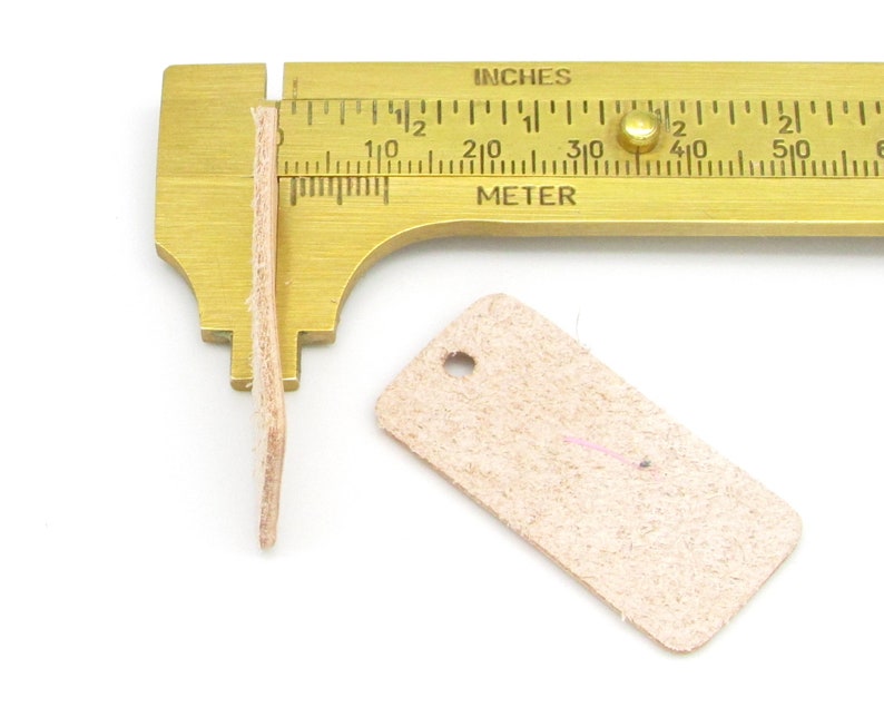 May include: A close-up of a leather tag with a hole in the top corner. The tag is being measured with a brass ruler, showing it is about 1.5 inches long.  The tag is a light beige color.