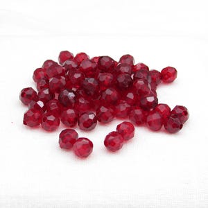 May include: A pile of faceted, deep red beads. The beads have a multi-faceted surface that catches the light, creating a sparkling effect. The beads are scattered on a white surface.