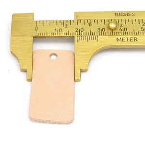 May include: A light brown leather tag with a hole punched in the top, measured with a ruler showing it is 1 inch wide and 1.5 inches long.