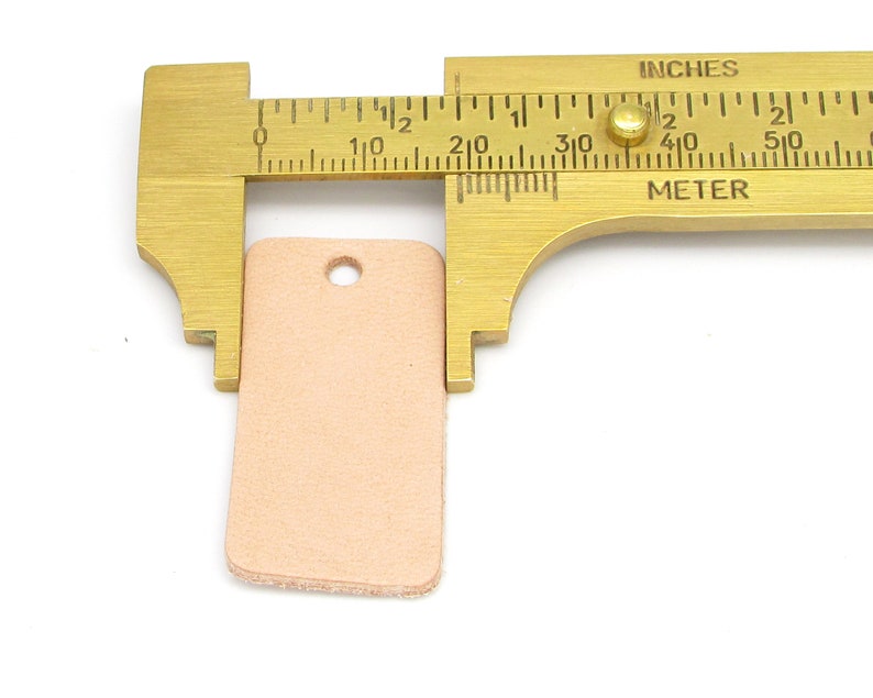 May include: A light brown leather tag with a hole punched in the top, measured with a ruler showing it is 1 inch wide and 1.5 inches long.