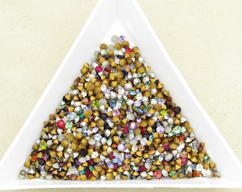 May include: A white triangular tray filled with a variety of small, colorful rhinestones. The rhinestones have a gold-colored base and are topped with various colors, including red, blue, green, and purple. The tray is on a light beige surface.