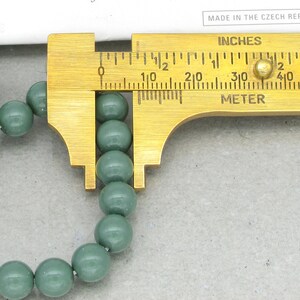 Pu&ograve; includere: Primo piano di una collana di perline rotonde verde scuro misurate con un calibro in ottone. Il calibro indica pollici e centimetri.