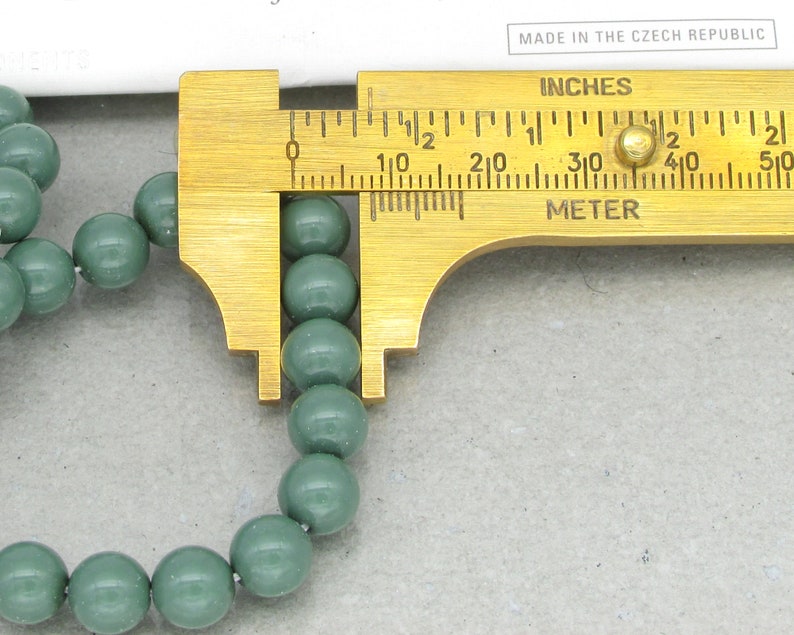 Pu&ograve; includere: Primo piano di una collana di perline rotonde verde scuro misurate con un calibro in ottone. Il calibro indica pollici e centimetri.