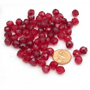 May include: A collection of faceted, translucent red beads. The beads are scattered on a white surface, with a U.S. penny for size comparison. The beads have a geometric shape and a glossy finish, suitable for jewelry making.