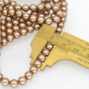 May include: A strand of brown pearl beads with a ruler measuring the size of the beads at 6mm. The beads are round and have a smooth, glossy finish.