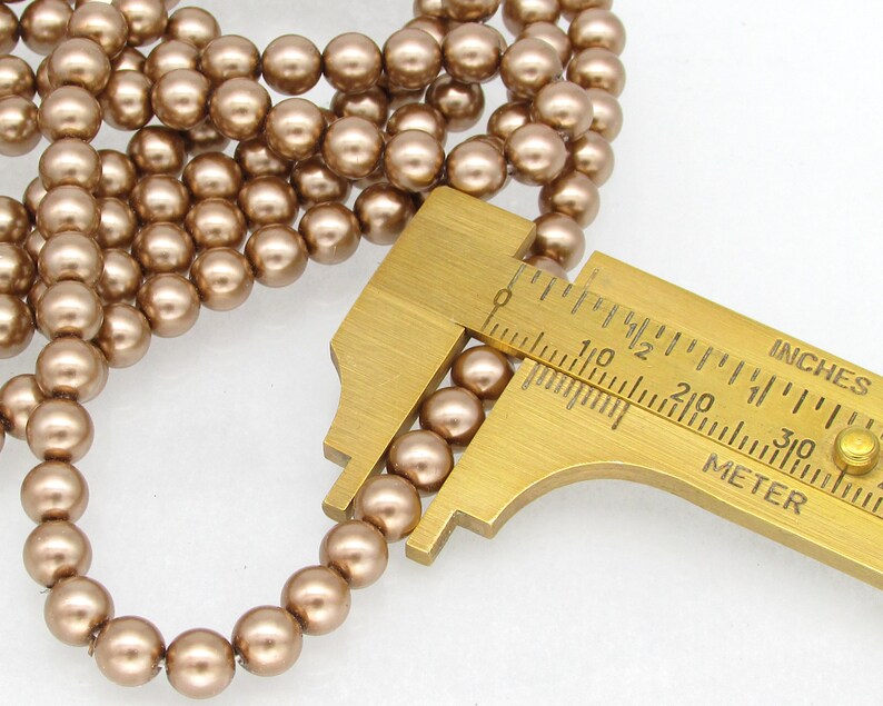 May include: A strand of brown pearl beads with a ruler measuring the size of the beads at 6mm. The beads are round and have a smooth, glossy finish.