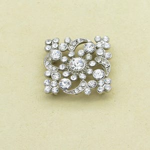 May include: A square, silver-toned brooch with a floral design. The brooch features numerous clear, round rhinestones of varying sizes, arranged in a symmetrical pattern. The design includes flower-like clusters and curved accents.