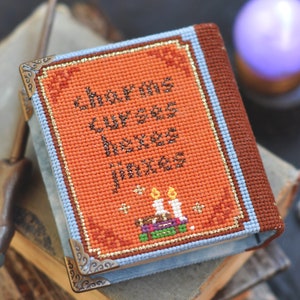 Pu&ograve; includere: Un piccolo libro arancione e marrone con il titolo "Charms, Curses, Hexes, Jinxes" ricamato sulla copertina. Il libro &egrave; decorato con bordi dorati e ha un bordo blu. Ci sono tre candele e una pila di libri sulla copertina.