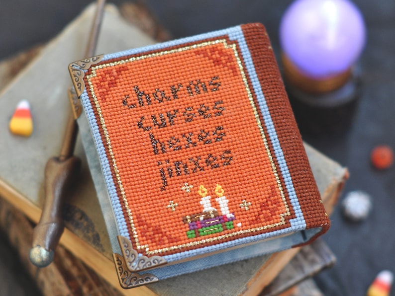 Pu&ograve; includere: Un piccolo libro arancione e marrone con il titolo "Charms, Curses, Hexes, Jinxes" ricamato sulla copertina. Il libro &egrave; decorato con bordi dorati e ha un bordo blu. Ci sono tre candele e una pila di libri sulla copertina.