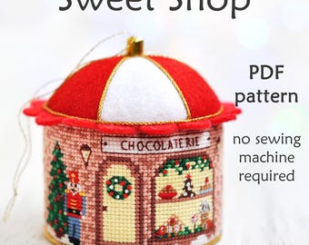 May include: A red and white felt Christmas ornament shaped like a small shop with a sign that reads "Chocolaterie". The ornament features a nutcracker, a Christmas tree, and a wreath. The text "PDF pattern no sewing machine required" is also included in the image.