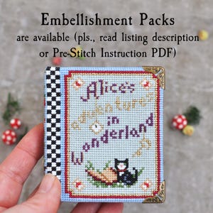 May include: A small, rectangular cross-stitch project featuring the text Alice's Adventures in Wonderland, a black cat, and a key. The design has a light blue background, a black and white checkered spine, and decorative gold-toned metal corner accents.