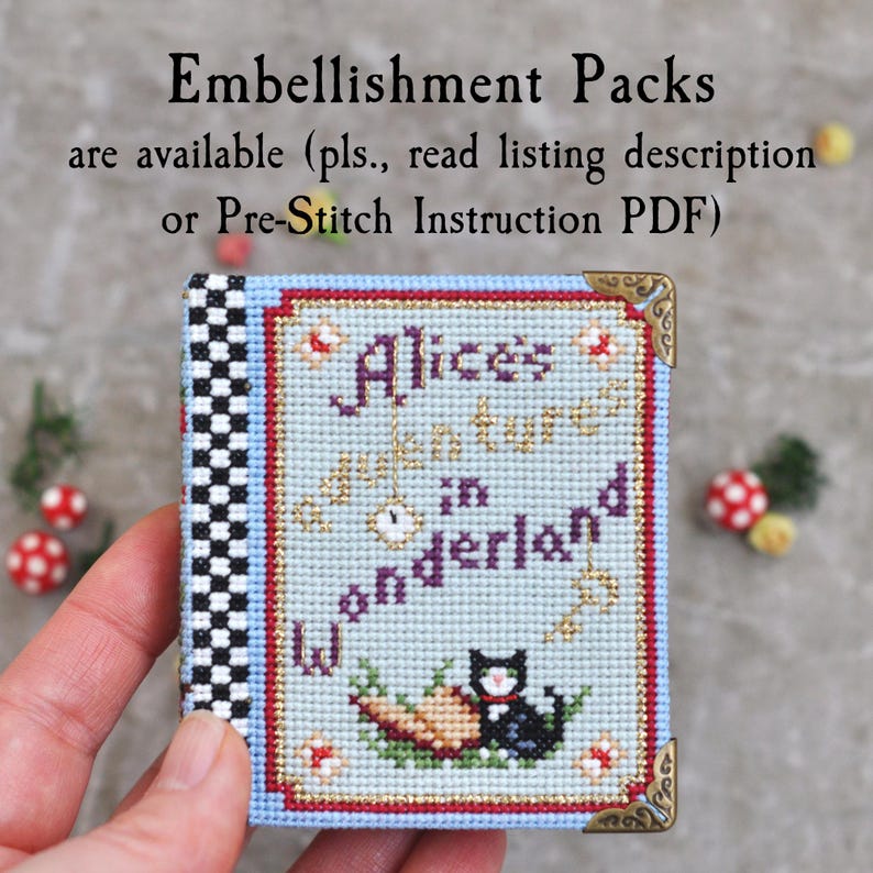 May include: A small, rectangular cross-stitch project featuring the text Alice's Adventures in Wonderland, a black cat, and a key. The design has a light blue background, a black and white checkered spine, and decorative gold-toned metal corner accents.