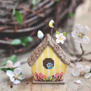 May include: A yellow and brown birdhouse pincushion with a bird, two eggs, and flowers on top. The pincushion is made of fabric and has a yellow and white striped design.