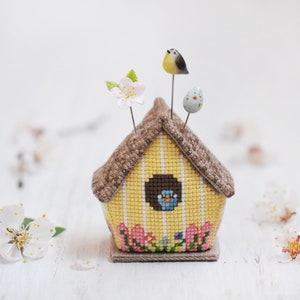 May include: A yellow and brown birdhouse pincushion with a blue bird peeking out of the window. The pincushion has a brown roof and a yellow base with pink flowers. There are three pins with a white flower, a blue egg, and a yellow bird on top.