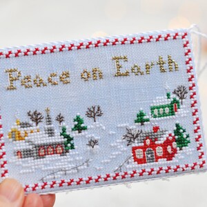 May include: A white cross-stitch ornament with a red and white border. The ornament features a winter scene with houses, trees, and the text "Peace on Earth" in gold thread.