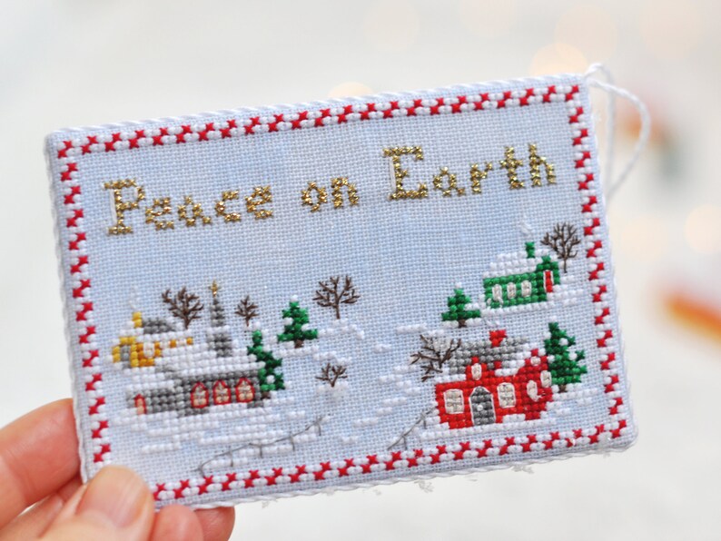 May include: A white cross-stitch ornament with a red and white border. The ornament features a winter scene with houses, trees, and the text "Peace on Earth" in gold thread.