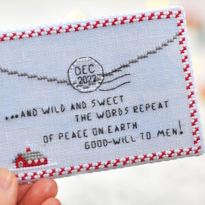 May include: A white cross-stitch ornament with a red border and a message that reads "...AND WILD AND SWEET THE WORDS REPEAT OF PEACE ON EARTH GOOD-WILL TO MEN!" with a red and white stamp that reads "DEC 2022".