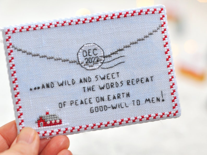 May include: A white cross-stitch ornament with a red border and a message that reads "...AND WILD AND SWEET THE WORDS REPEAT OF PEACE ON EARTH GOOD-WILL TO MEN!" with a red and white stamp that reads "DEC 2022".