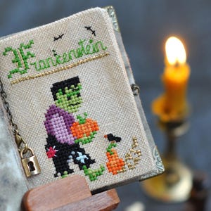 May include: A cross-stitched book cover with the word "Frankenstein" in green and gold thread. The cover features a pixelated Frankenstein's monster holding a pumpkin. A small lock and chain secure the book. A lit candle is in the background.