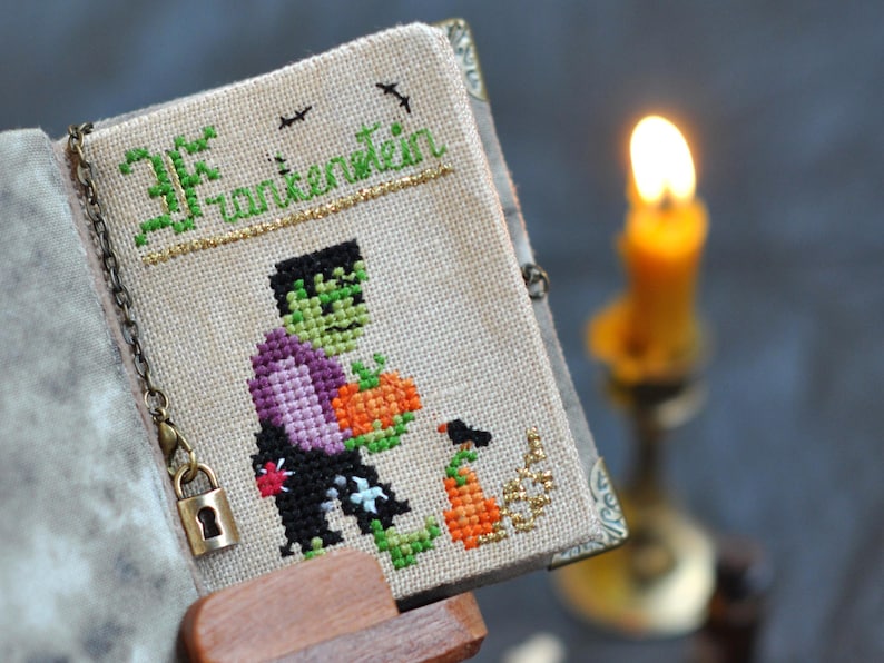 May include: A cross-stitched book cover with the word "Frankenstein" in green and gold thread. The cover features a pixelated Frankenstein's monster holding a pumpkin. A small lock and chain secure the book. A lit candle is in the background.