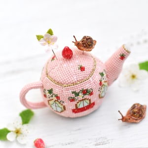 May include: A pink crocheted teapot pincushion with a strawberry design and a snail on top. The teapot has a handle and a spout. There are two other snails on the white surface.