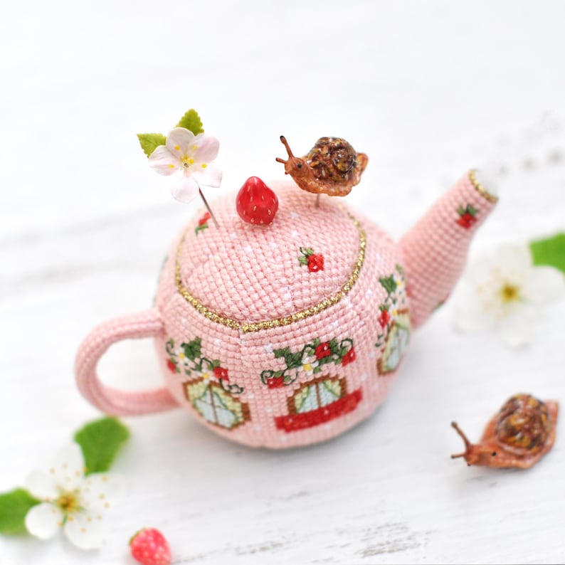 May include: A pink crocheted teapot pincushion with a strawberry design and a snail on top. The teapot has a handle and a spout. There are two other snails on the white surface.