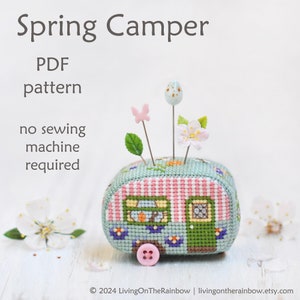 May include: A cross-stitched camper-shaped pin cushion with decorative pins. The camper is light blue with pink and green accents, a pink button wheel, and the text "Spring Camper PDF pattern no sewing machine required". Decorative pins include a butterfly, egg, and flower.