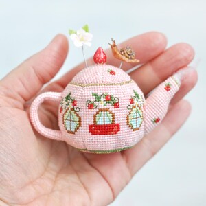 May include: A pink teapot pincushion with a strawberry on top, decorated with green leaves, red strawberries, and windows. A small snail is perched on the top of the teapot.