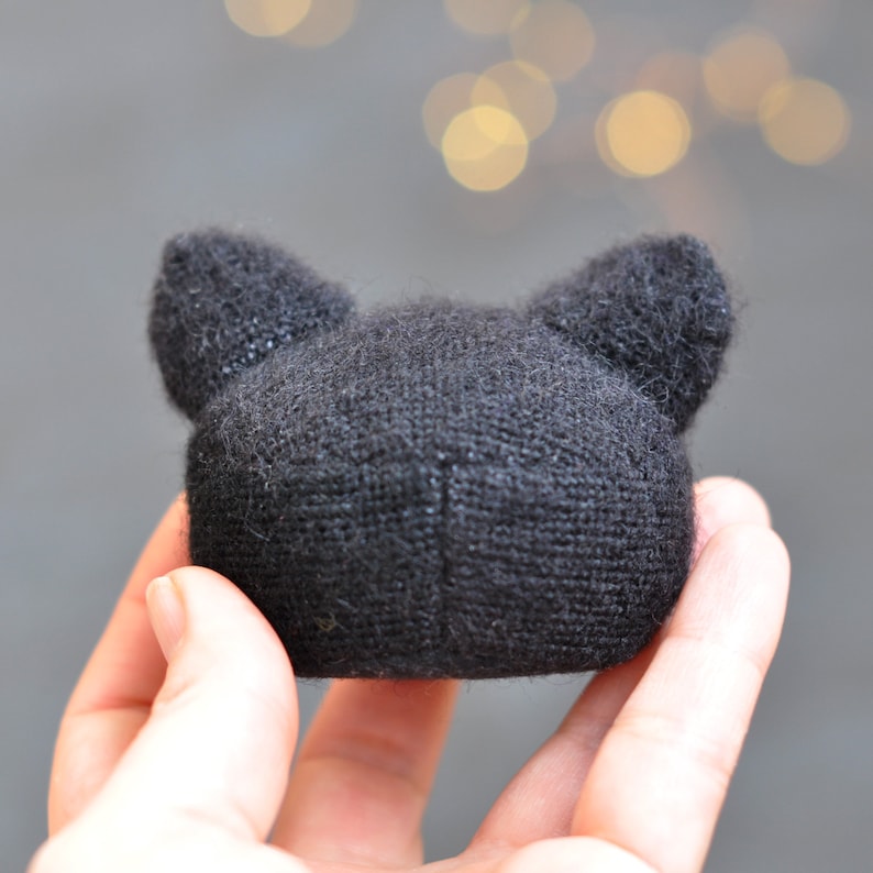 May include: A black knitted cat head with large ears. The cat head is soft and plush.